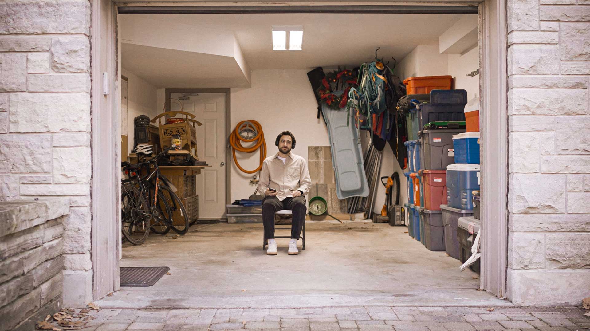 man sits in a cluttered garage, wearing headphonesman sits in a cluttered garage, wearing headphones listening to a podcast.