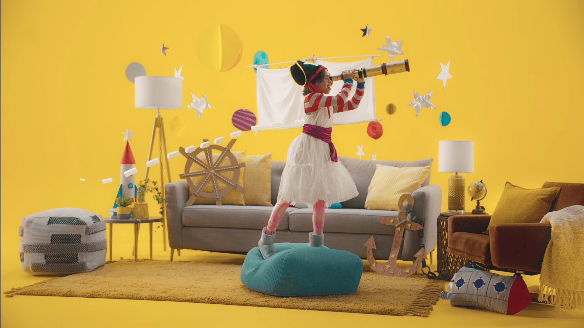 A child dressed as a pirate plays with a telescope in a bright living room filled with imaginative decor, including stars and ships, against a vibrant yellow background.