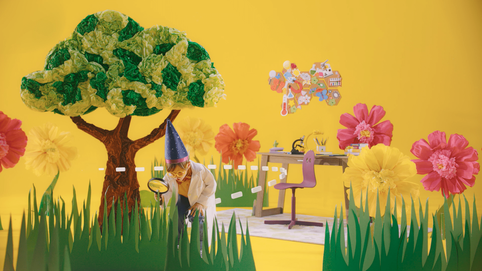 A child in a wizard costume investigates with a magnifying glass near a large, colorful paper tree and flowers in a playful, science-themed room