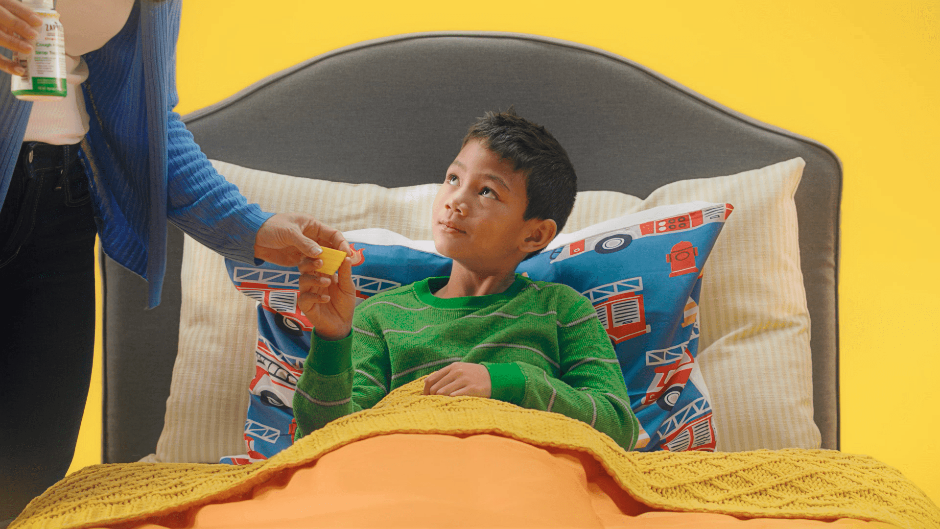 A child resting in bed receives a dose of Zarbee's syrup from an adult hand while gazing up from under cozy blankets